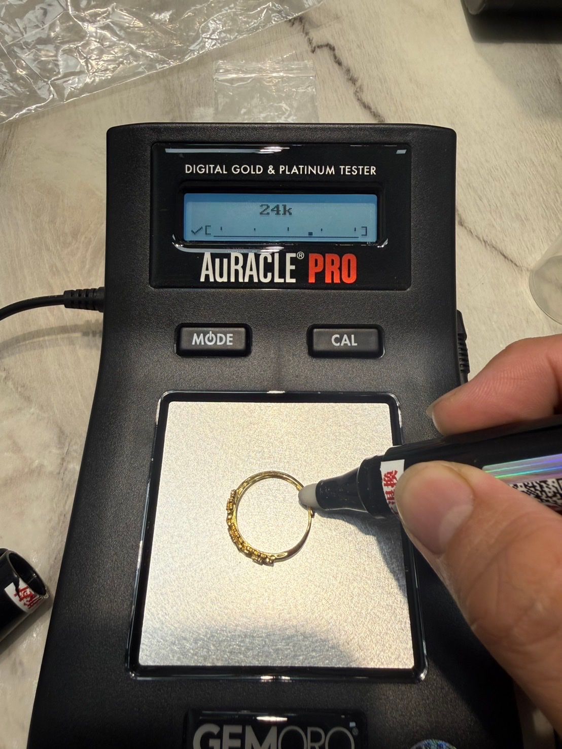 店內以 AuRACLE PRO 黃金測試儀檢測金飾外層成色，螢幕顯示 24k